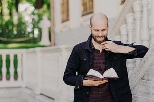 Clever middle aged student learns and cramms information before final exam, focused in book, has gentle smile, thick beard, poses outdoor near old building. People, leisure and education concept photo