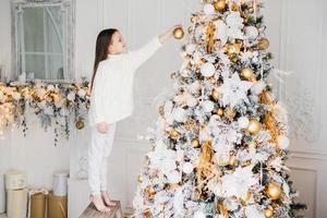 Indoor shot of small kid in white clothes, decorates New Year tree, stands on tiptoe, holds decoration in hands, does everything without parents help. House decorating and childhood concept. photo