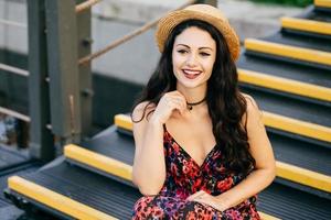 Smiling female with dark luxurious hair wearing dress and straw hat sitting at stairs looking with delightful look into distance. Restful young woman with appealing appearance sitting outdoors photo