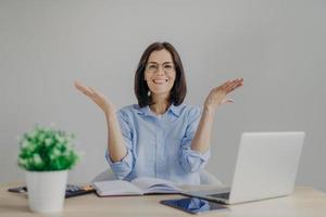 Happy overjoyed female achieves success in her work, going to present high results, raises hands joyfully, sits at working place with laptop computer, diary, calculator, isolated on grey background photo