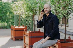 Attractive bald male with beard, has pleasant talk with girlfriend via cell phone, spends free time outdoor in park, being in good mood. Positive smiling young businessman has rest during weekends photo