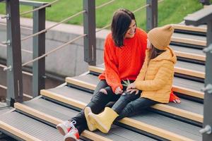 la imagen de una joven de moda usa un suéter rojo, mira positivamente a un niño pequeño, posan juntos en los pasos al aire libre, disfrutan del tiempo libre, pasean por el entorno urbano. personas, concepto de relación foto