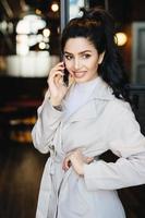 Beautiful young woman with dark hair tied in pony tail and nice eyes dressed in white raincoat speaking over smartphone holding hand on waist while standing outside looking down having happy look photo