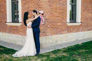 Horizontal portrait of romantic bride and bridegroom embrace as stand outdoor near ancient builduing at green grass, look at each others eyes, express their love and romance. Marriage concept photo