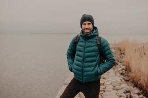 Horizontal shot of attractive unshaven man has cheerful expression, wears hat and jacket, keeps hands in pocket, carries rucksack, has walk during foggy autumn weather, poses near lake outdoor photo