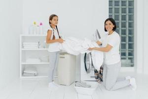 Glad housewife does washing with little adorable helper. Mother and daughter wash clothes in laundry room, load linen in washer. Woman stands on knees near washing machine. Housework concept photo