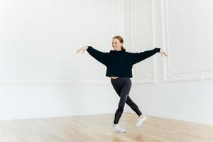 Professional female ballerina practices dance in hall, stretches hands, stands with crossed legs, has gentle smile on face, dressed in black sportswear, enjoys spare time for hobby. Healthy lifestyle photo