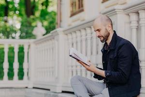 Sideways shot of bearded male with bald, holds book, sits outdoor against white balcony background, reads interesting detective story or scientific literature. People, education and hobby concept photo