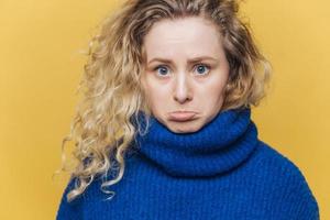 Attractive offended young female has curly bushy hair, curves lower lip in dissatisfaction, feels upset after quarrel with husband, wears blue sweater, isolated over yellow studio background. photo