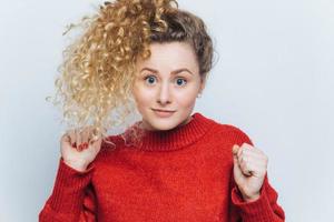 Horizontal shot of beautiful blue eyed funny female with curly bushy hair tied in pony tail, keeps hands pressed in fists, raises them, has unexpected reaction on something shocking, isolated on white photo