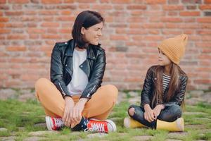 Shot of pleased realxed two sisters pose crossed legs, sit on lawn, pose against brick wall background, have happy facial expressions. Little kid in leather jacket spends time with her mother photo