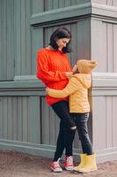 Vertical shot of attractive caring mother looks at eyes of her daughter with love, embrace while pose outside, wears ripped jeans, sweater and sneakers, smile positively. Family relationship concept photo