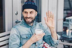 Horizontal portrait of happy male with dark beard and mustache, waves with hand, glad to notice friend, holds cup of tea, wears fashionable clothing. Man greets someone. Hipster guy drinks coffee photo