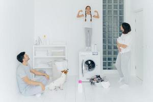 Photo of happy schoolgirl poses on top of washing machine, shows muscles, raises arms, ready to help parents with washing or laundry. Woman and man pose in washing room with dog and small kid