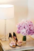 Beautiful shoes with high heels, lamp and bouquet with pink flowers standing on bedside-table. Bouquet and shoes of bride. Wedding shoes and flowers. Preparation for special occasion