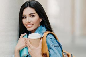 foto al aire libre de una estudiante morena con maquillaje, cabello largo oscuro, vestida con una chaqueta de mezclilla, toma café para llevar, libro de texto, lleva una mochila, mira a algún lugar a lo lejos, pasea por la ciudad