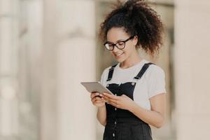 Pretty curly haired girl looks at digital tablet screen with smile, reads some text or electronic book, wears spectacles for vision correction, white t shirt and dungarees, enjoys high speed internet photo