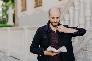 Handsome clever bald male holds book, reads interesting novel with serious expression, stands outdoor against ancient white building, likes reading. People, hobby, lifestyle and leisure concept photo