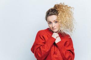 Horizontal portrait of pleasant looking young female with curly light pony tail, dressed in red sweater, looks with non confident expression at camera, poses against white wall with blank copy space photo