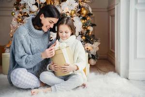 Indoor shot of adorable girl and her mother sit crossed legs, open wrapped present box, have intrigue what is there, sit against adorned New Year or Christmas tree, have sincere gentle smiles on faces photo