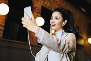 Brunette woman in white jacket having dark eyes with long eyelashes, full lips and healthy skin having good mood while resting in cafe listening to different tracks with earphones and making selfie photo