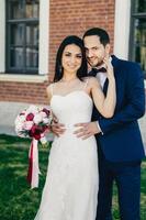 Pleasant looking young female brunette in white wedding dress holds bouquet stands near her handsome bridegroom who wears formal suit, stand outdoors, pose in camera, have happy expressions. photo