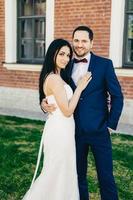 una pareja romántica enamorada celebra su boda, se abrazan cariñosamente, tienen expresiones felices, se paran al aire libre. bonita joven pareja de novios después de la ceremonia. gente, amor, concepto de matrimonio foto