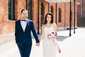 Bearded male bridegroom wears formal black suit keeps his brides hand, has happy expression, celebrate wedding together. Romantic couple in love have walk outdoors after ceremony. Love concept photo