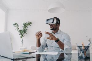 African american businessman use virtual reality glasses working at laptop at office desk. High-tech photo