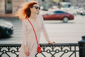 Horizontal shot of red haired woman wears sunglasses, focused aside, poses near hence at street, poses against road with transport, strolls in city during summer sunny day, copy space for your text photo