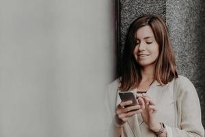 chica de aspecto agradable con corte de pelo a la moda, pasea afuera, envía mensajes por teléfono celular, disfruta de la conexión a Internet en roaming, usa una elegante chaqueta blanca, usa la aplicación, chatea con amigos foto