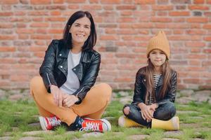 Horizontal shot of restful mother and daughter sit in lotus pose, wears fashionable leather jackets, sneaks and boots, sit on ground against brick wall, feel relaxed and carefree. Lifestyle, family photo
