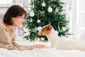 Horizontal shot of beautiful cheerful woman enjoys togetherness with dog, holds its paws, lie on white bed, look each other in eyes, feel love and friendship, decorated Christmas tree in background photo