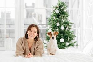 Cheerful brunette woman gets jack russell terrier dog as Christmas gift, lies on bed, feels relaxed and happy, green decorated New Year tree in background. People, animals, holidays concept. photo