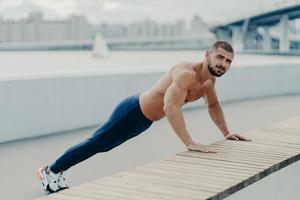 El hombre musculoso en forma hace ejercicio de empuje hacia arriba lleva un estilo de vida saludable usa pantalones deportivos y zapatillas de deporte posa al aire libre cerca del puente. deportista motivado empujando duro. deporte, motivación y determinación foto