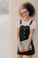 Vertical shot of cheerful young Afro student carries notepads or diary, wears white t shirt, black sarafan, ready for having classes, stands outdoor. People, studying and university concept. photo