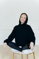 Relaxed cheerful woman in black sweatshirt, sits in lotus pose on chair, sits indoor, has glad expression, poses indoor, satisfied after active training with coach. People, rest, motivation concept photo