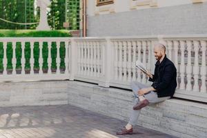 Shot of clever male professor reads scientfic literature, prepares for lectures at university, poses outdoor, holds book, sits crossed legs, wears fashionable clothes, has beard, enjoys calmness photo