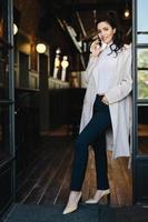 Stylish young female with dark hair wearing white coat, black trousers and shoes with heels posing into camera while standing against cafe background chatting over phone holding hand in pocket