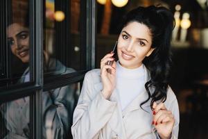 Cute female with wavy dark hair, charming eyes, well-shaped lips and white even teeth wearing white clothes having red manicure communicating over telephone touching her hair with her gentle hand photo