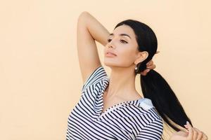 Sideways shot of good looking brunette woman keeps hair in pony tail, looks confidently into distance, thinks about something, shows her luxurious hair, stands indoor against light studio wall photo