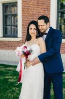 Newlyweds express their love, stand together outdoors. Loving couple celebrate their wedding. Beautiful female bride in white long dress and bouquet stands back to his affectionate bridegroom photo