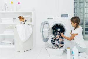 Busy child does laundry work, empties washing machine, cleaned clothes in basin uses detergents, little pedigree dog in basket. Modern household device at home. Female kid helps with family chores photo