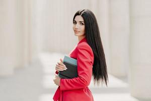 Serious brunette businesswoman with makeup, manicure, wears red jacket, carries diary and notepad, stands in profile outdoor, goes for work. Elegant businesslady on street. Stylish director outside photo