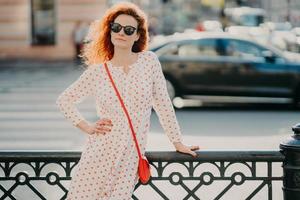 Outdoor shot of satisfied redhead woman keeps one hand on waist, other on street hence, poses over blurred urban setting, wears sunglasses and dress, has red manicure. Copy space aside for text photo