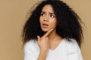 Puzzled female model keeps fore finger on chin, looks thoughtfully aside, has frustrated facial expression, curly Afro hair, wears casual white jumper, isolated over brown background. Close up shot photo