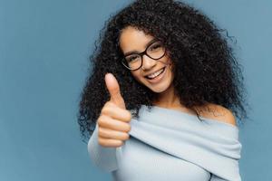 Its excellent Glad smiling African American woman shows like gesture, keeps thumb up, gives approval, cheers over blue wall, dressed in casual clothes, expresses approval of amazing product photo