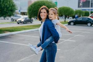 Image of affectionate mother gives piggyback to small daughter, smile broadly, have outdoor walk, pose against blurred background with cars, play together. People, family and happiness concept photo