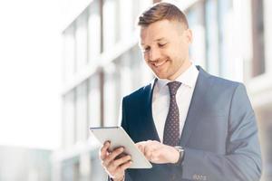 Handsome prosperous businessman reads useful information in internet via touchpad, connected to wireless internet, poses outdoor. Confident office worker makes booking online. Technology concept photo