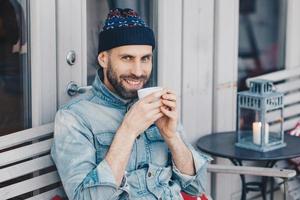 Portrait of handsome male with cheerful expression, enjoys spare time, drinks coffee or tea, wears hat and denim jacket, being in good mood. Attractive man has thick beard and mustache poses indoor photo